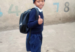 Zdjęcie przedstawia kenińskie dziecko w niebieskim mundurku z plecakiem szkolnym na plecach